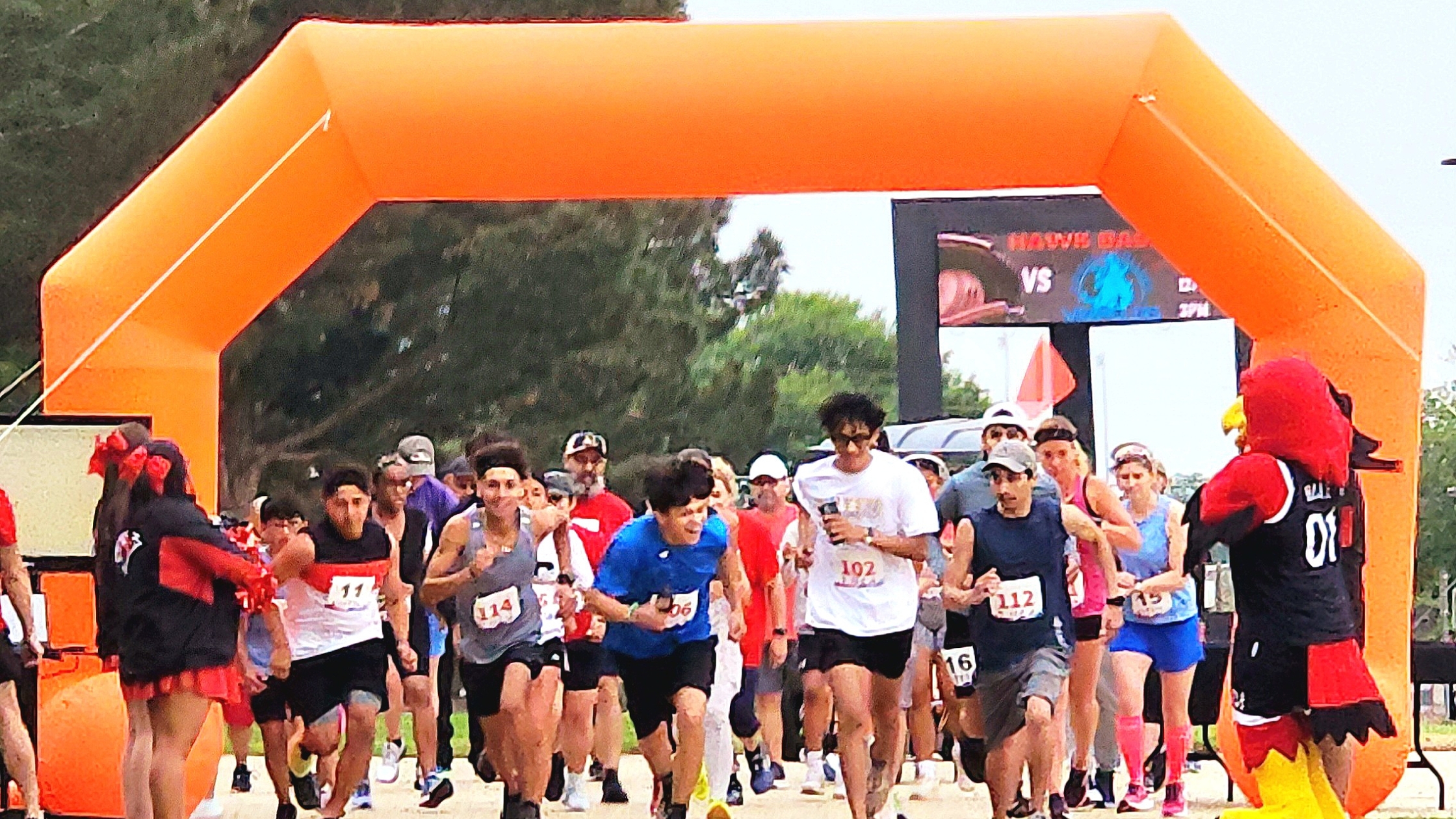 Runners take off from the starting line under a large orange inflatable arch at the beginning of a 5K race. Participants wear numbered bibs and athletic gear, with a mix of serious and excited expressions. On either side of the arch, Howard College cheerleaders and the school mascot, a red hawk in a black jersey labeled '01,' cheer on the runners. A digital scoreboard is visible in the background, and trees line the course.
