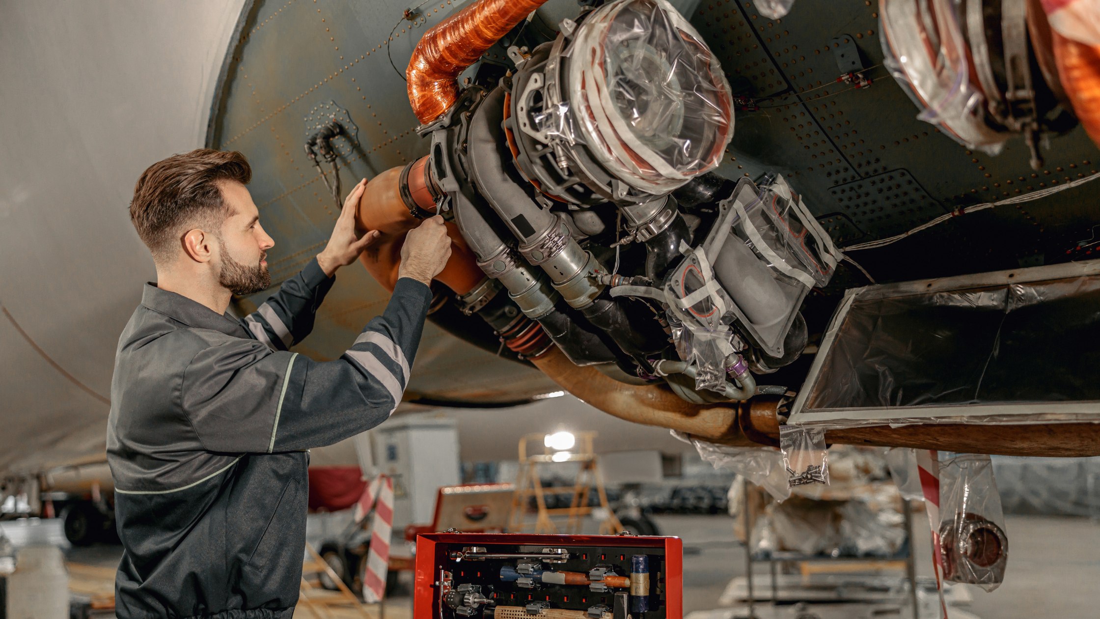 A male technician works on the engine of an aircraft in a hangar, surrounded by tools and equipment. Image used to promote a new aviation maintenance program.