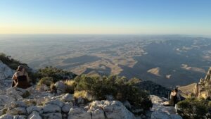 Two hikers sit on rocky terrain near the summit of Guadalupe Peak, overlooking a vast desert landscape stretching to the horizon. The scene is bathed in soft evening light, highlighting the ridges and valleys below under a clear blue sky.