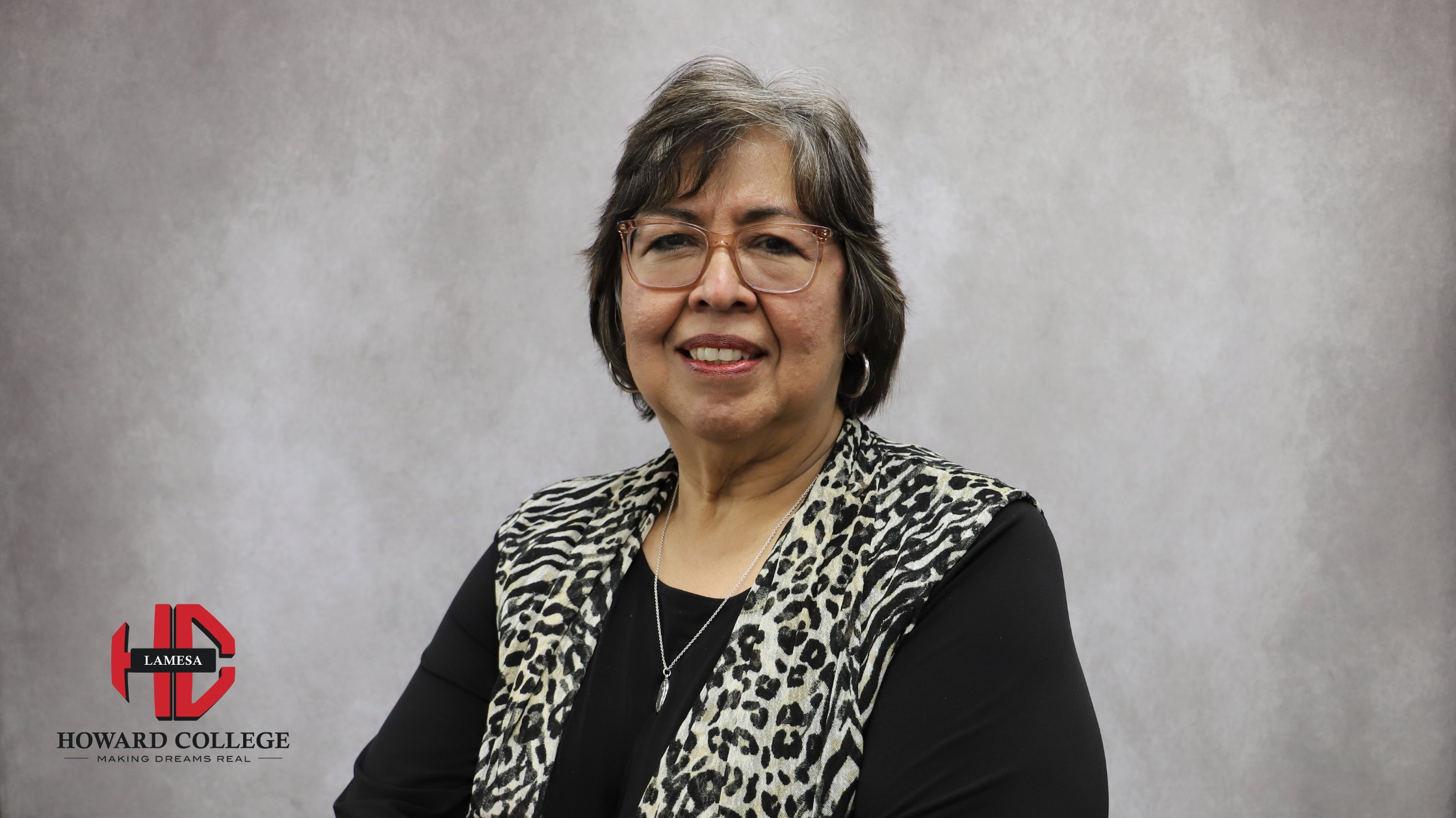 Portrait of a smiling woman with short gray hair, wearing glasses, a black top, and a black-and-white animal print vest, posed in front of a gray background with the Howard College Lamesa logo.