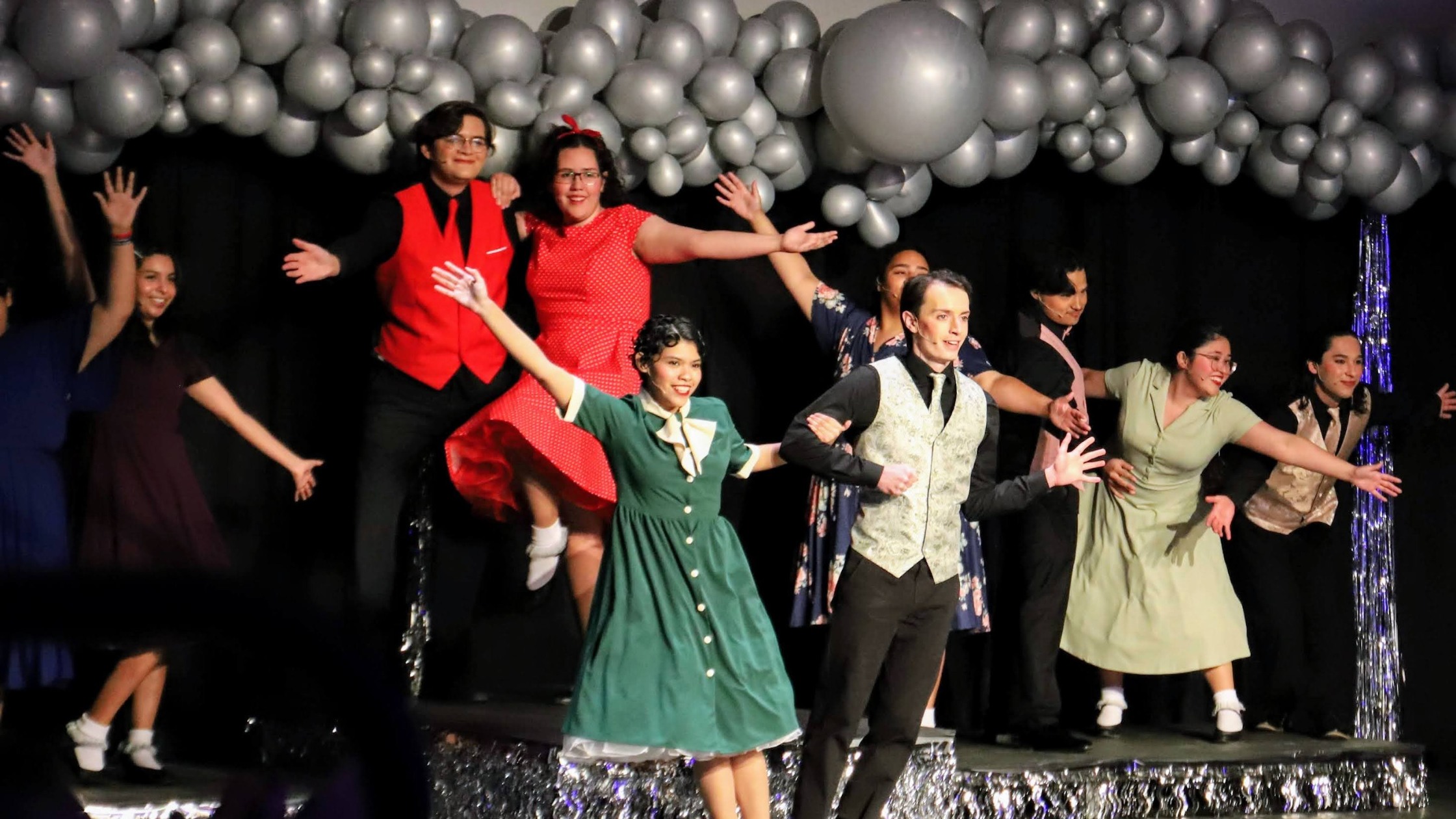 Students perform a lively musical number on stage dressed in colorful vintage costumes, surrounded by silver balloons and festive decorations.